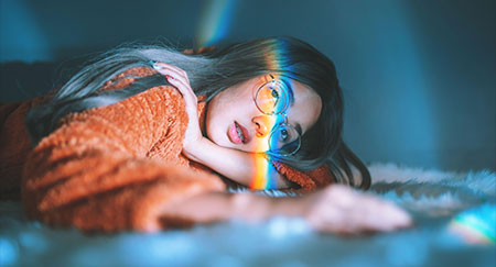 Femme allongée sur un lit avec une lumière aux couleurs de l’arc-en-ciel sur le visage.