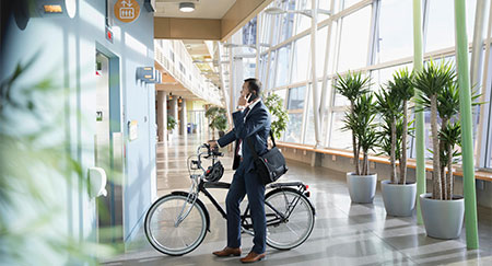 Businessman commuter with bicycle talking on smart phone, waiting at elevator in office lobby.