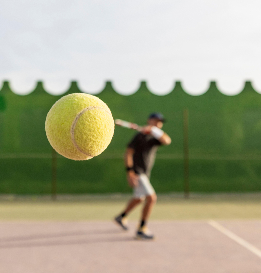 Tennisspieler, aufgenommen mitten in der Bewegung bei einem Rückhandschlag, wobei der Fokus auf dem Tennisball liegt und der Spieler im Hintergrund zu sehen ist