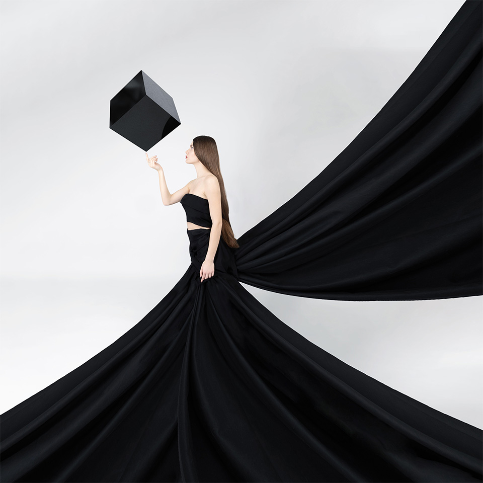 Mode, beaux-arts, concept fantastique. Magnifique femme aux longs cheveux bruns avec une très grande robe noire et une traîne qui vole comme des ailes, touchant un gros cube noir en lévitation.