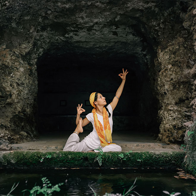 Barfuß sitzende Frau mit geschlossenen Augen meditiert in Lotussitz in der Nähe einer Steinhöhle."