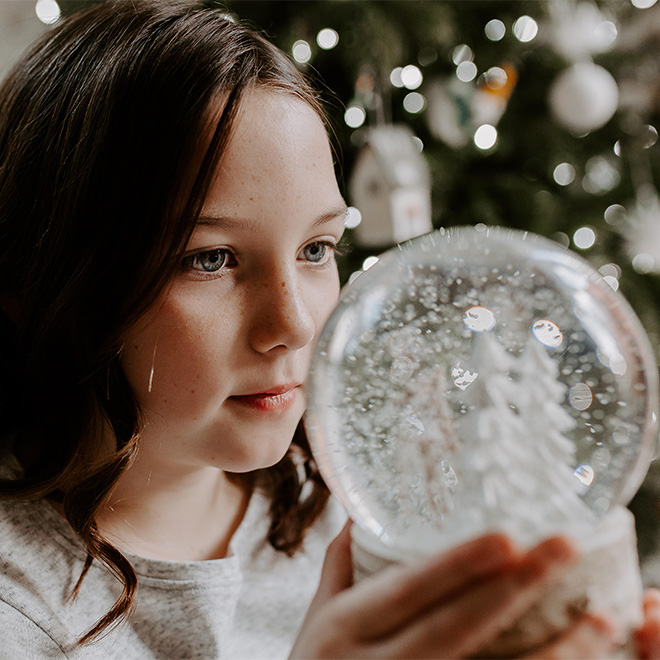 Mädchen sitzt neben einem Weihnachtsbaum und betrachtet eine Schneekugel.