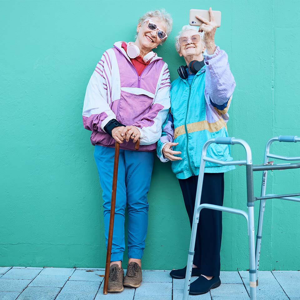 Telefono, selfie e disabilità con amiche anziane in posa per una fotografia all'aperto con una parete verde come sfondo. Felicità, cellulare, deambulatore e anziani.