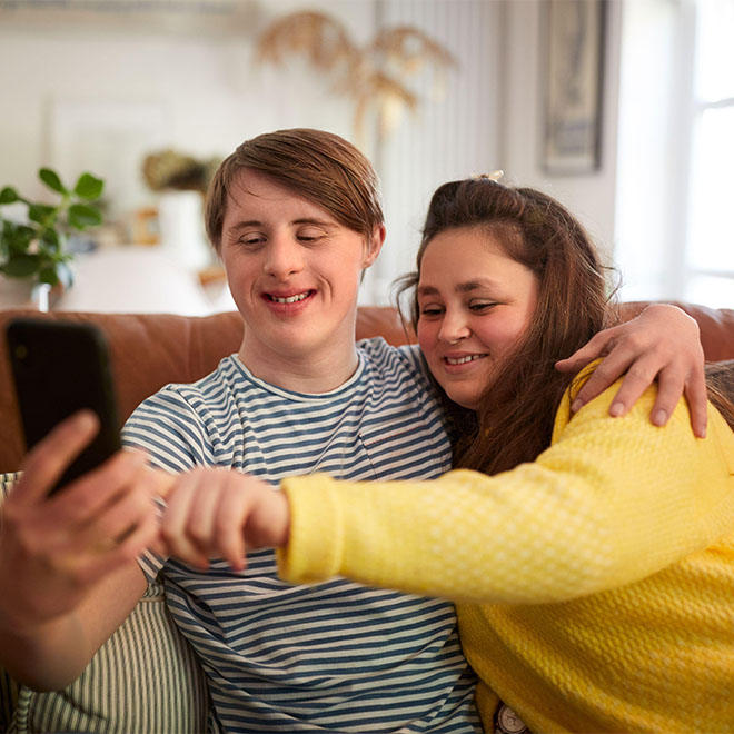 Junges Paar mit Down-Syndrom sitzt auf dem Sofa und benutzt ein Mobiltelefon, um zu Hause ein Selfie zu machen.
