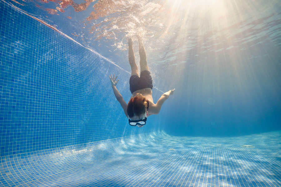 Ganzkörperaufnahme eines barfüßigen Jungen mit Schwimmbrille und Shorts, der unter der Sonne in einem sauberen Pool schwimmt