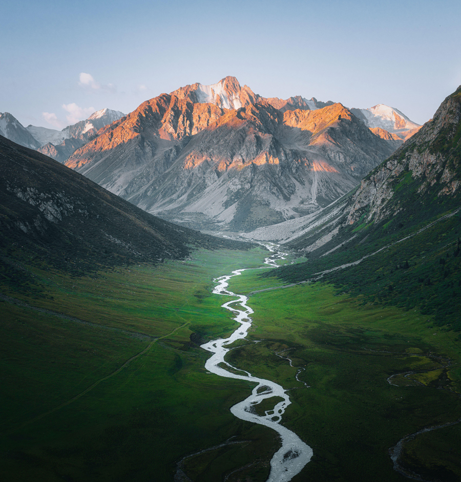 Aerial view of a winding river snaking through a vibrant green valley, framed by rugged mountains kissed with golden light, Karakol, Issyk-Kul Region, Kyrgyzstan