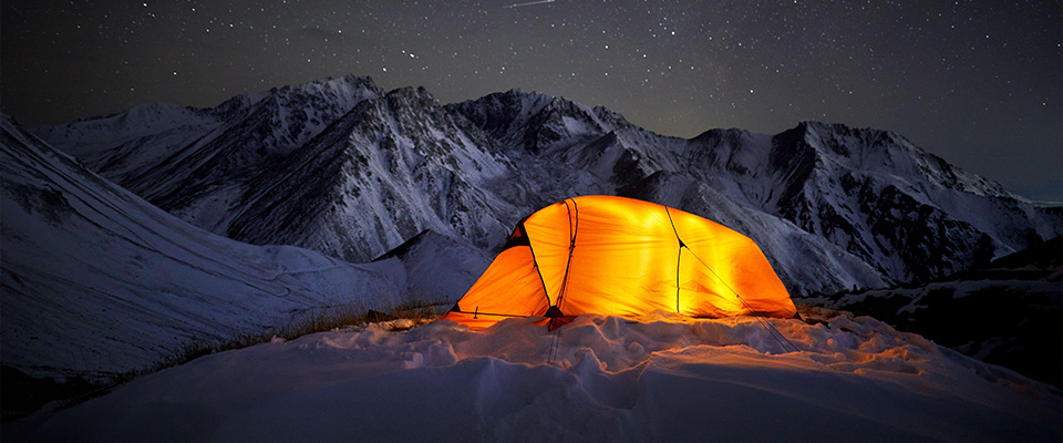 2DTR4EH – Leuchtendes oranges Zelt in winterlicher Berglandschaft im Dunkeln mit nächtlichem Himmel und Sternschnuppe im Hintergrund