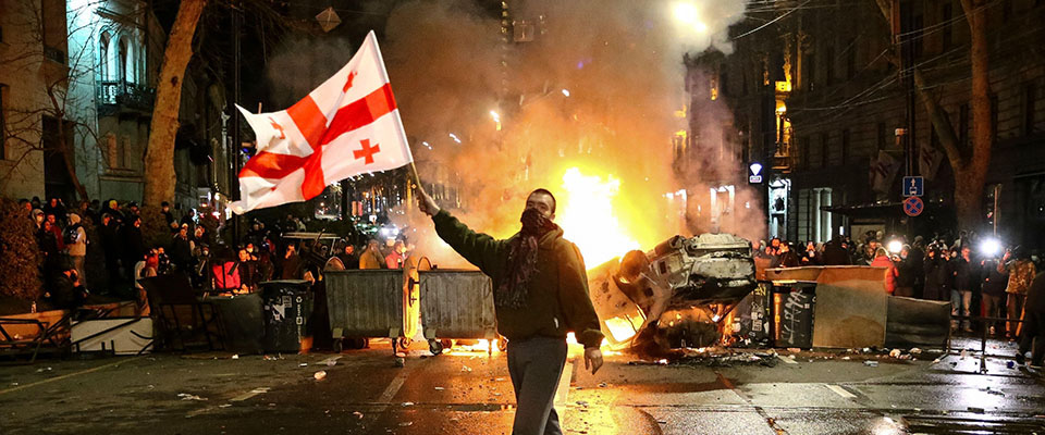A man waves a Georgian national flag in front of a burning barricade as other protesters stand behind not far from the Georgian parliament building in Tbilisi, Georgia, Thursday, March 9, 2023.