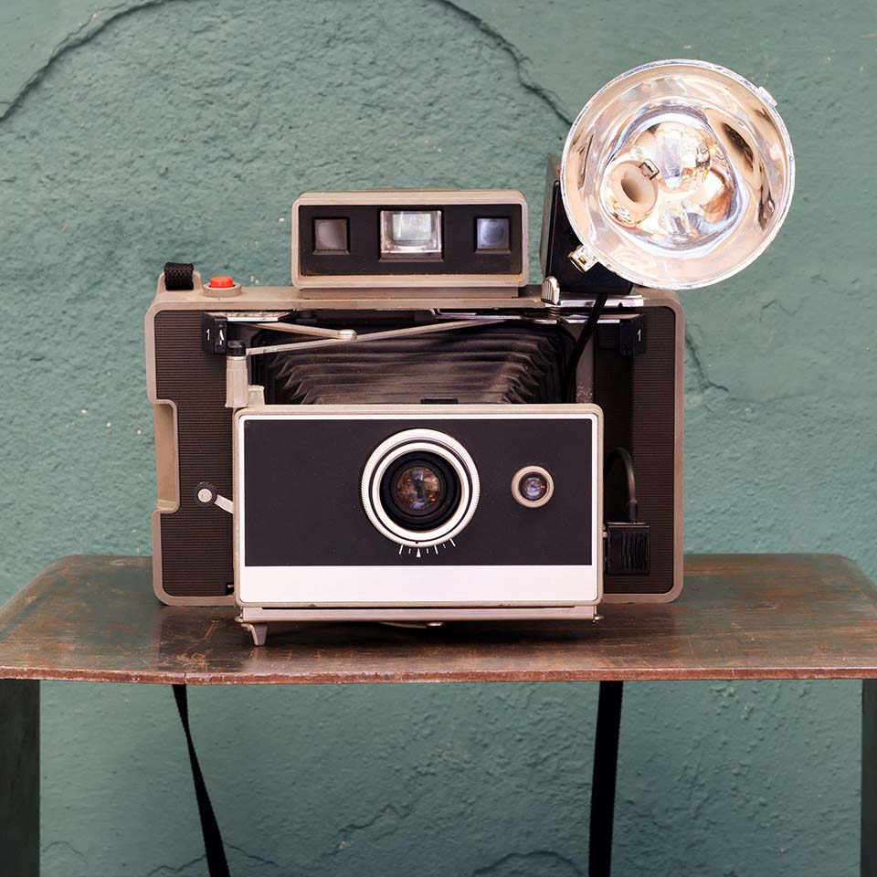 2C97CMM - Appareil photo instantané vintage avec soufflets et lampe sur une petite étagère devant un mur vert texturé, abîmé dans un concept photographique