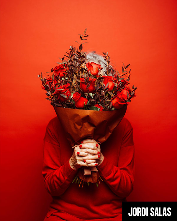 Senior woman wearing red clothes, hidding her face with a red roses bouquet, over a red background