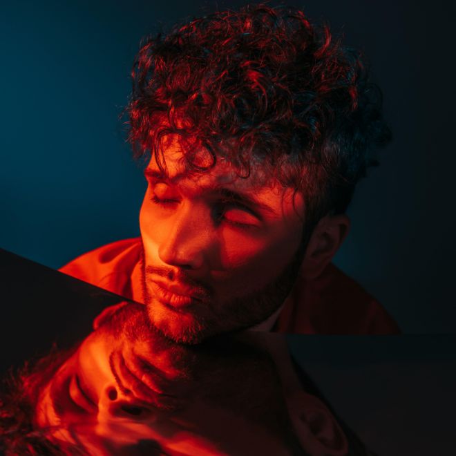 curly and stylish man in orange shirt posing with closed eyes near mirror on blue - Image ID: 2JJ1YW0 (RF)