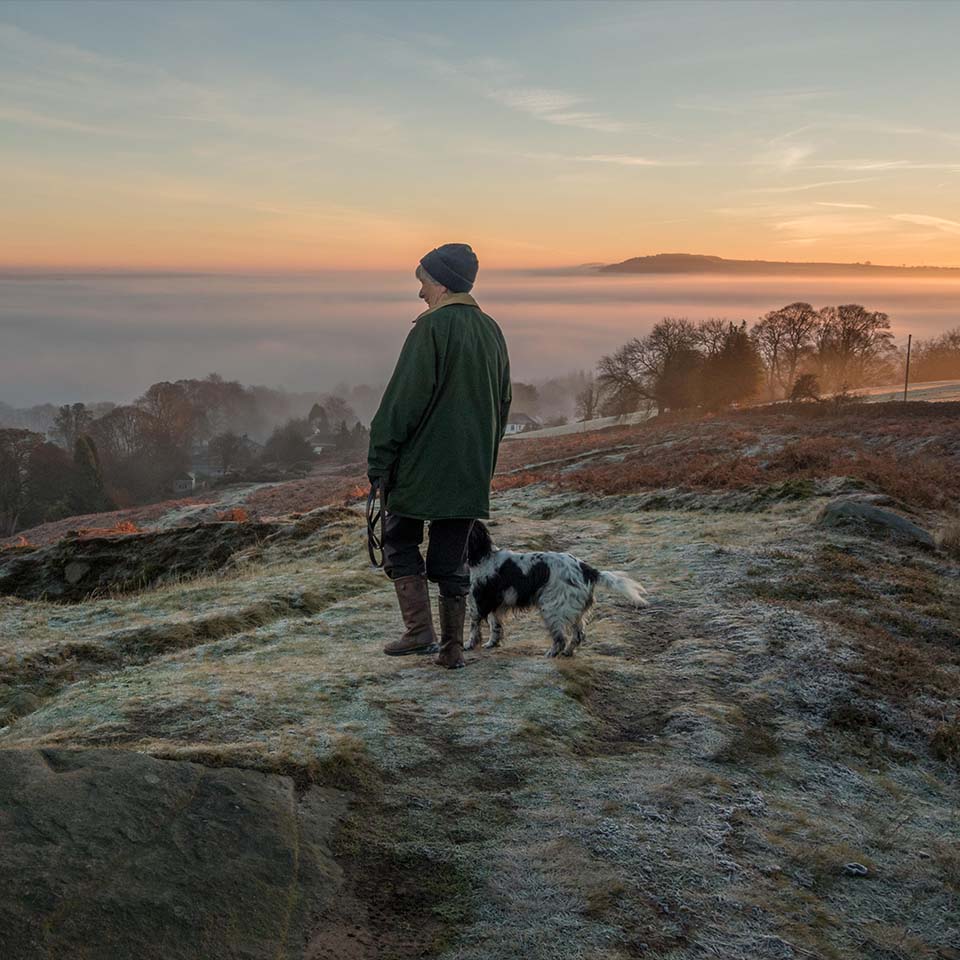 J67671 – Frau, die früh an einem frostigen Morgen mit ihrem Hund auf einem Pfad im Ilkley Moor spazieren geht und in die Landschaft blickt, während sich am Himmel Wolken bilden, Wharfe Valley, Yorkshire, Großbritannien