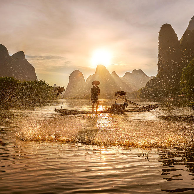 Ein Kormoran-Fischer wirft sein Netz auf dem Li-Fluss in Xingping, Guilin, China, während die Sonne hinter den Karstbergen hinter ihm untergeht.