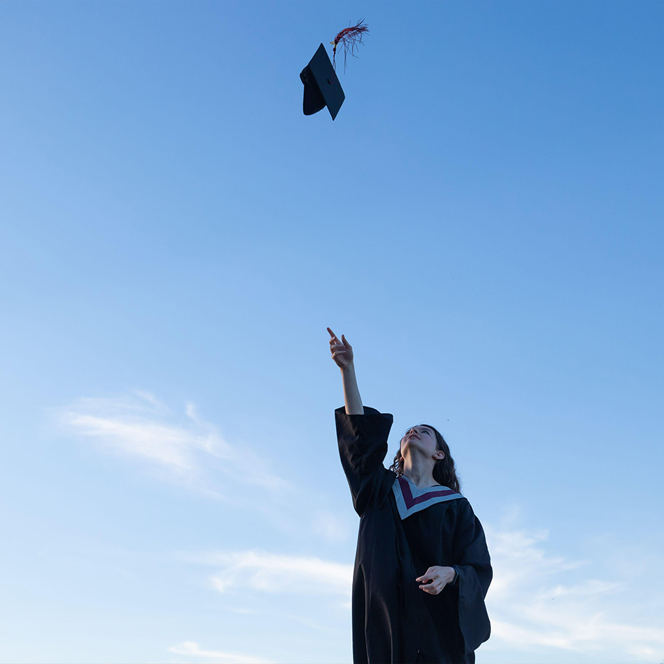 Young woman wearing graduation gown tossing her cap, mortar board, into the air with a blue sky in the background.