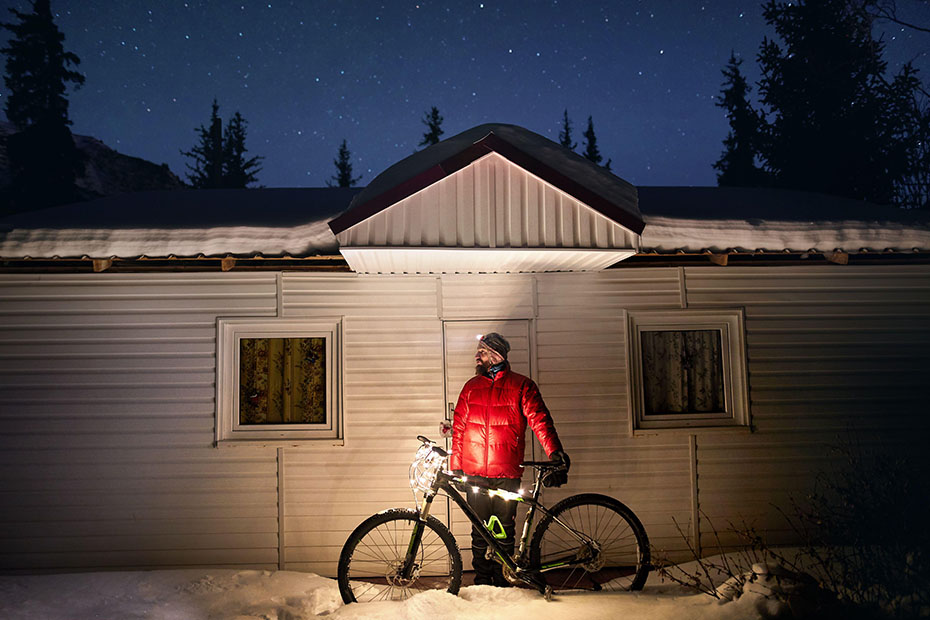 Man in red jacket with bicycle decorated with Christmas lights near small house at winter snowy forest in the mountains under night sky with stars 
