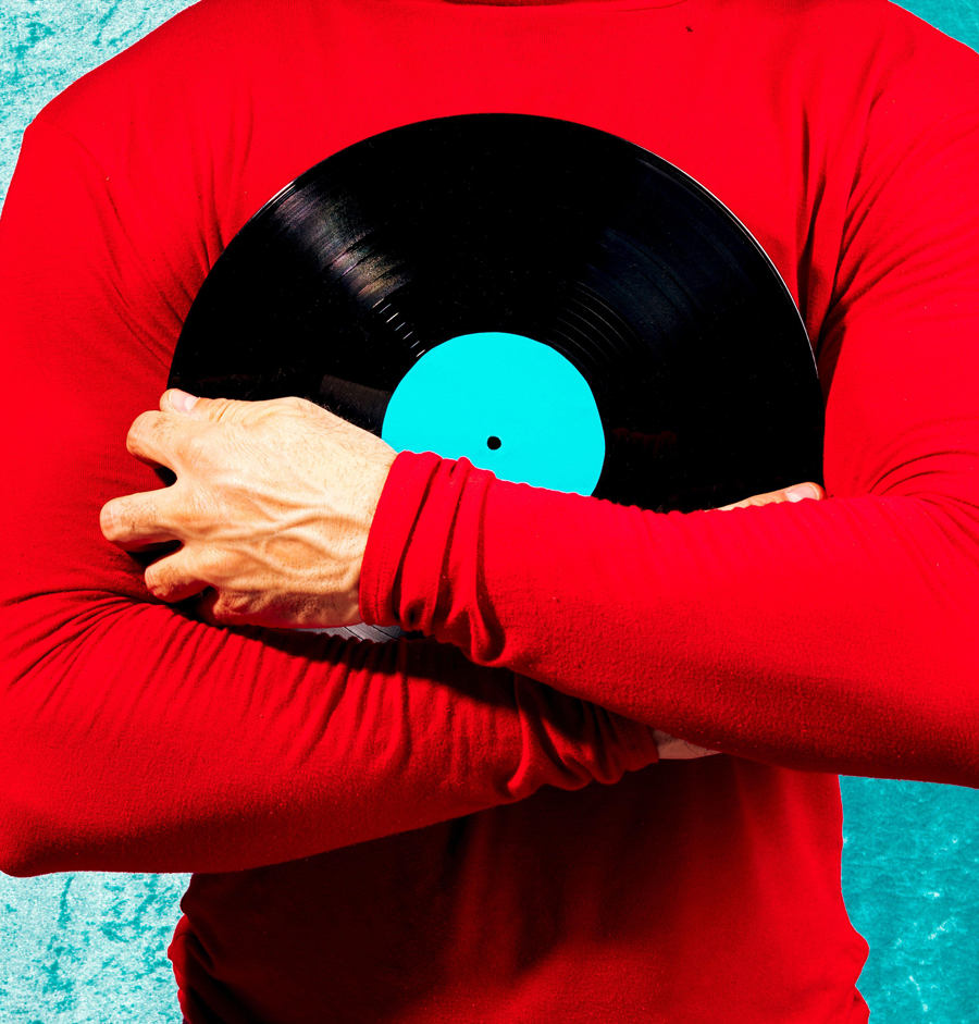 Cropped view of a person in a red shirt holding a black vinyl record with blue label against a turquoise backdrop