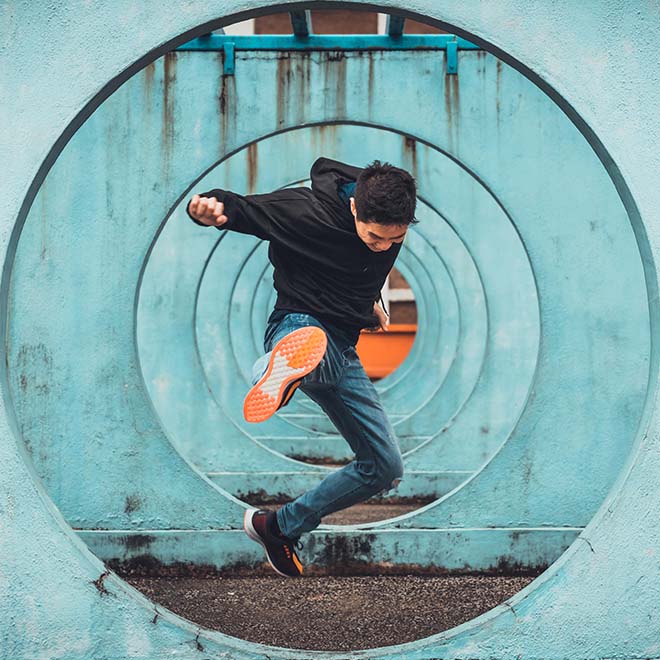 Young male student kicking in the air whilst shot through multiple concrete circle structures. 