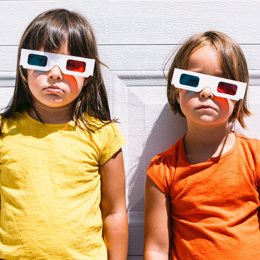 Two girls in casual colorful clothes and three dimensional glasses standing on white wall background looking at camera