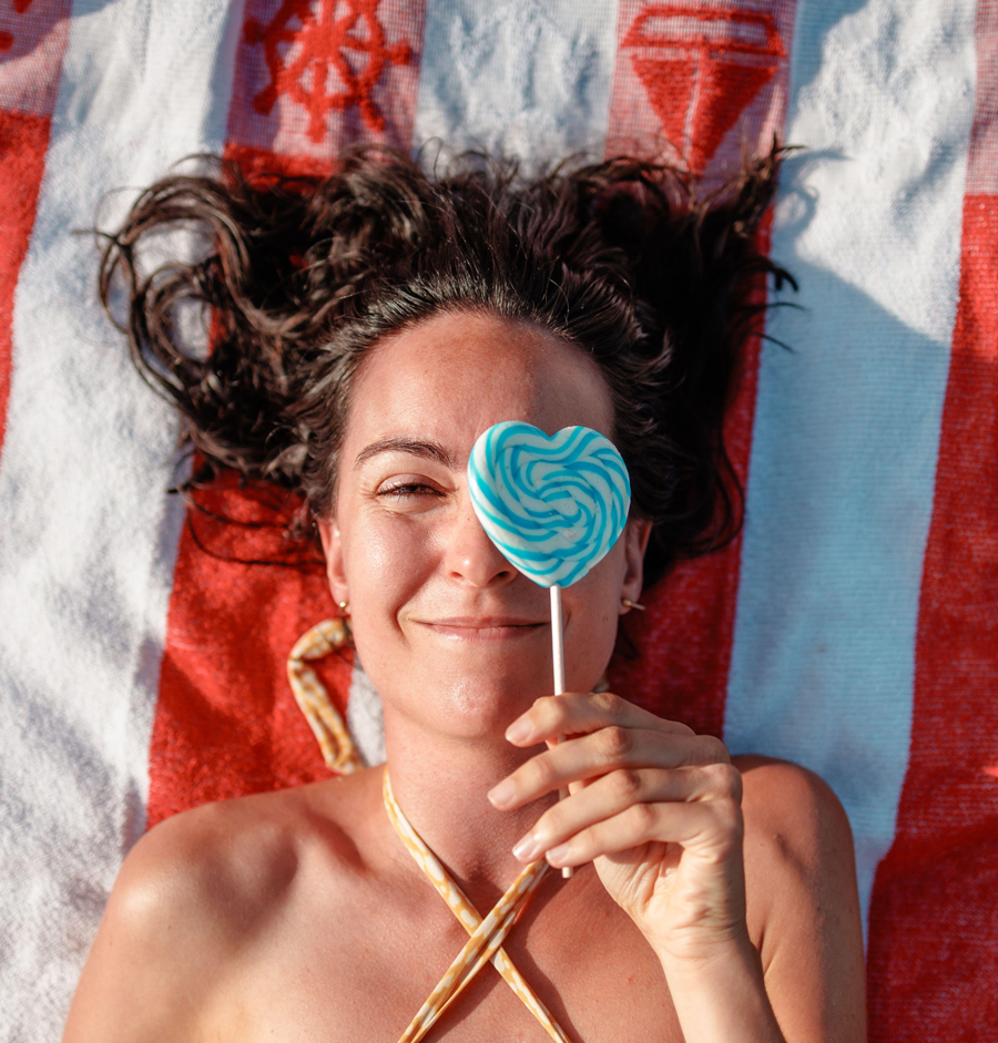 Smiling woman lying on a striped towel by a lake in summer, holding a blue heart-shaped lollipop over one eye, enjoying the sunshine