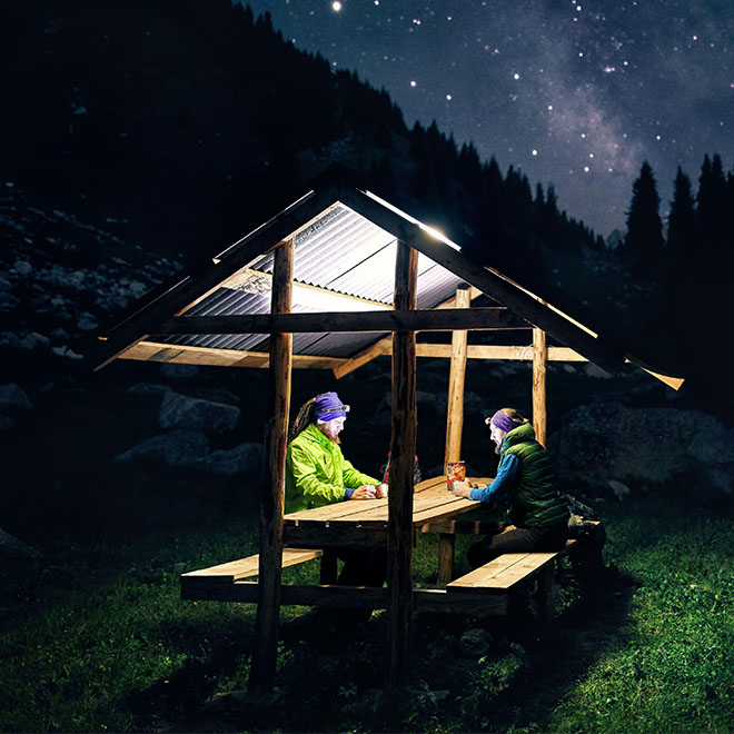 Zwei Männer sitzen in einem Sommerhaus in den Bergen unter einem Nachthimmel mit Sternen und der Milchstraße.