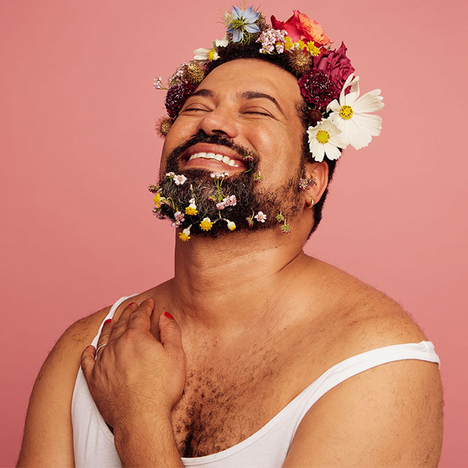 Genderqueer sorride su sfondo rosa. Uomo gay truccato con fiori sulla testa e sulla barba con spazio per il testo."