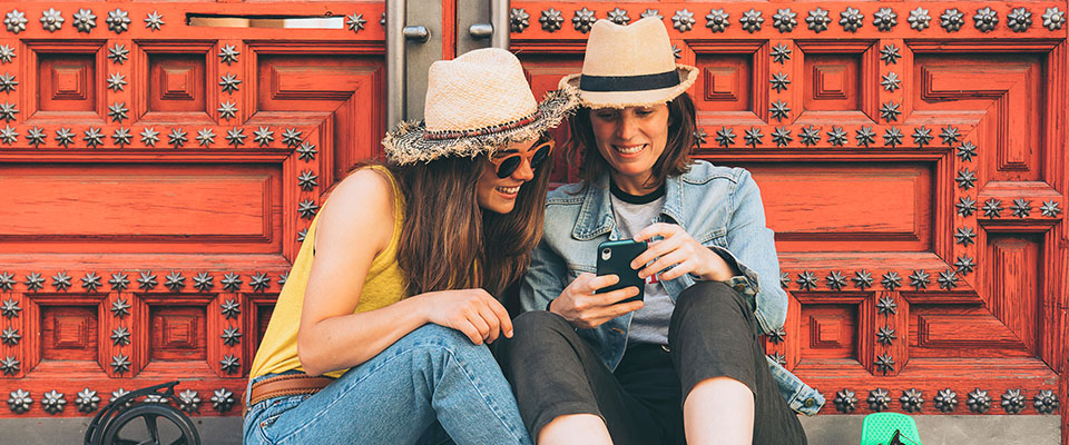 T8JE3H - Attraente coppia di giovani donne lesbiche guardano il telefono cellulare e sorridono l'un l'altra su uno sfondo di porta rossa. Concetto di felicità e gioia tra persone dello stesso sesso. 