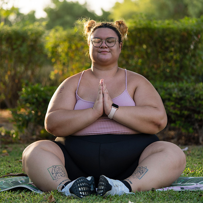 Plus-size Frau sitzt auf einer Yoga-Matte im Freien und meditiert lächelnd.