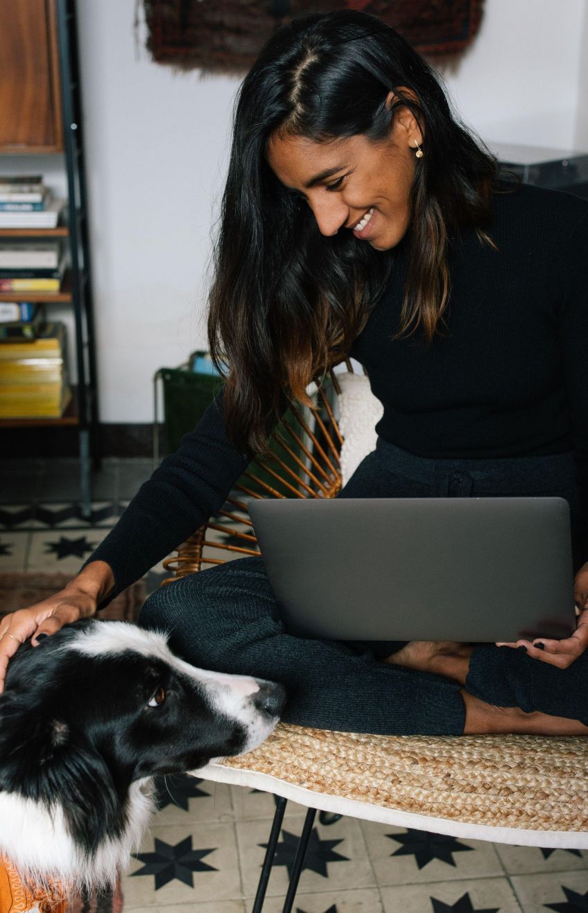 Delighted Indian female freelancer sitting on chair and working remotely from home with obedient Border Collie dog sitting nearby