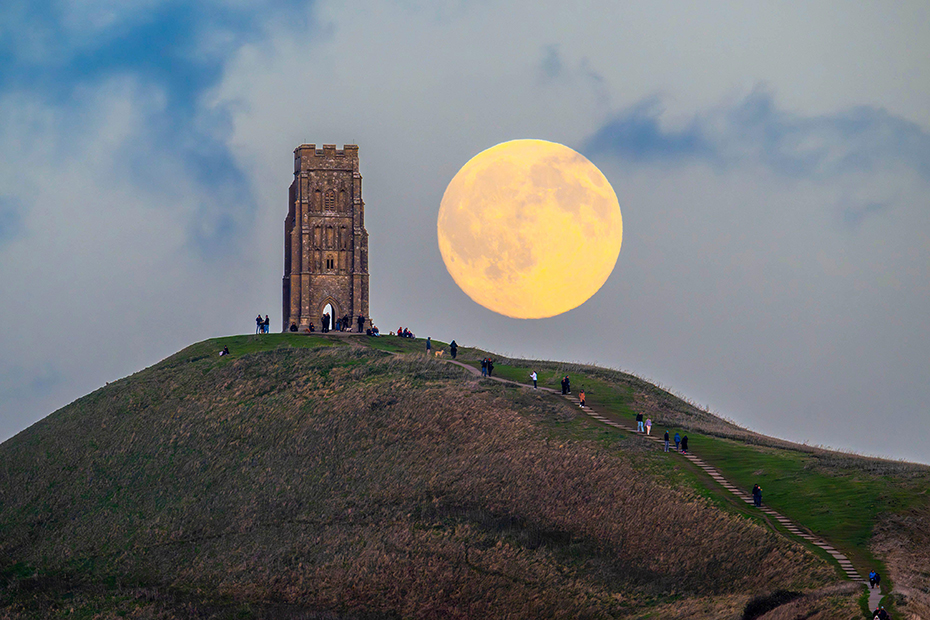 The almost full Hunter’s Moon rises up into the evening sky from behind St Michael’s Tower on Glastonbury Tor in Somerset as a crowd of people gather on top of the hill to watch. 