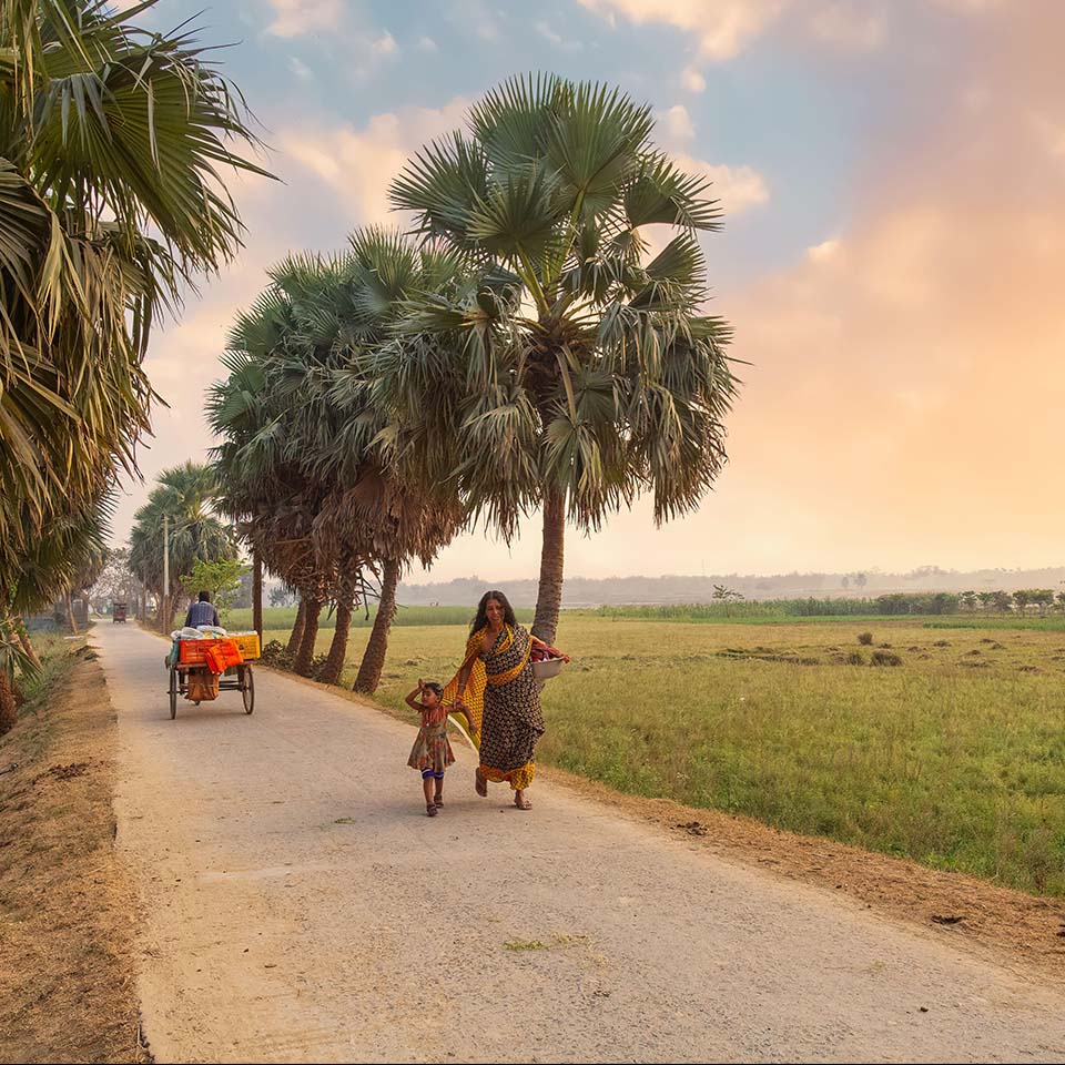 2EWTP9F – Ländliche Frau mit ihrem Kind auf einer unbefestigten Dorfstraße mit Blick auf landwirtschaftliche Nutzflächen in einem Bezirk in Westbengalen, Indien