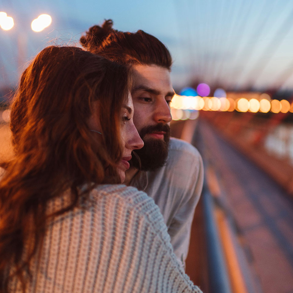 Nachdenkliches Paar schaut weg, während es auf einer Brücke in der Stadt in der Abenddämmerung steht.