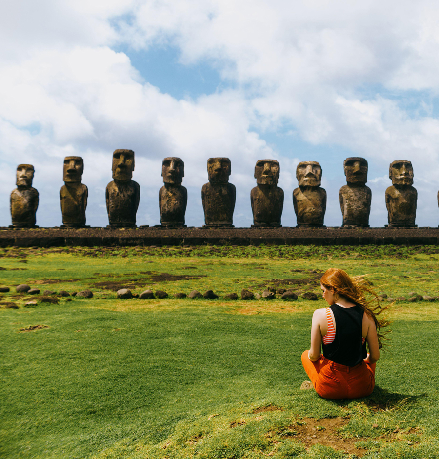 A person with flowing long hair sits on the grassy field in front of the iconic Moai statues of Easter Island