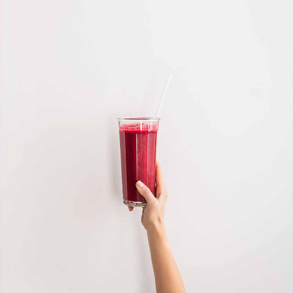 Main humaine tenant un verre de smoothie frais à la betterave et à la grenade violette avec une paille en verre sur un fond de mur blanc, espace copie. Concept de détox, de régime, de perte de poids, de mode de vie sain.