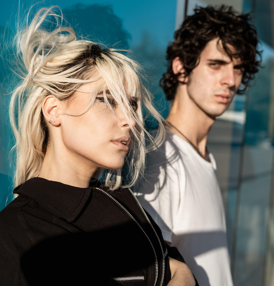 Young man and woman looking towards camera while standing against blue wall with reflection of street in sunlight