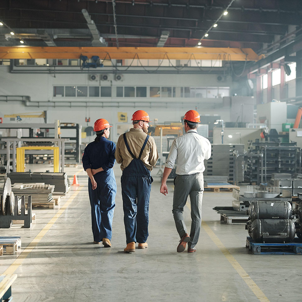 2B12BP1 – Rückenansicht von drei Arbeitern einer Industrieanlage, die an einer großen Fabrik entlang gehen