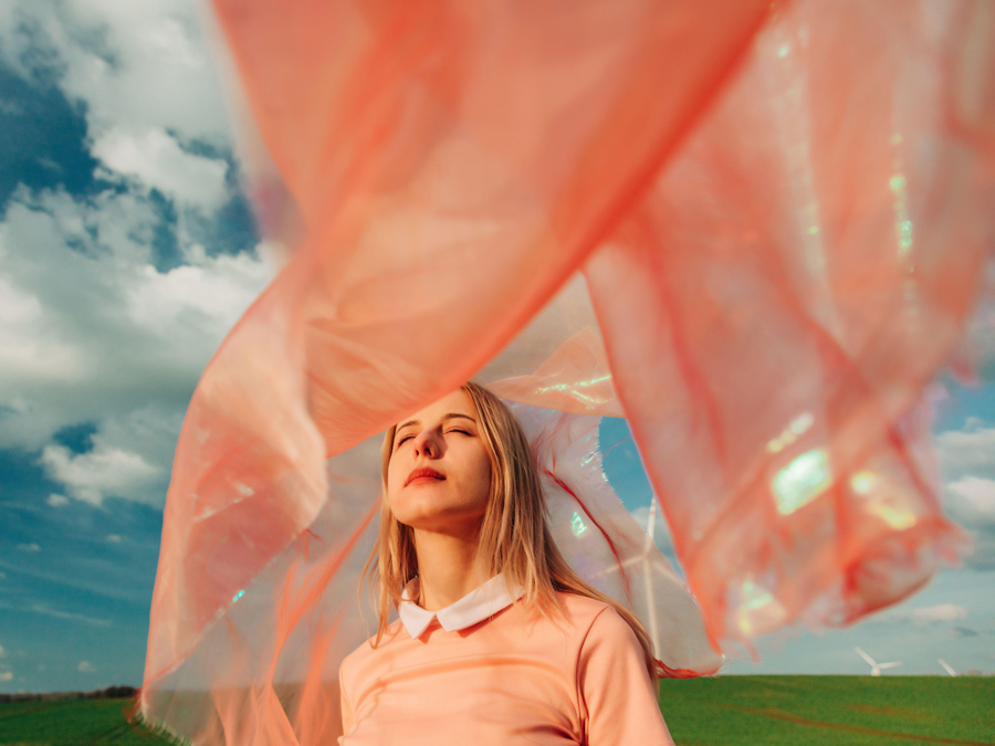 A woman stands in a field with her eyes closed, enveloped by a flowing pink fabric