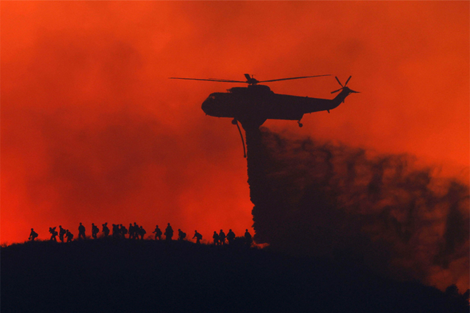 A water tanker drops water while firefighters walk below, as the Sandy Fire approaches in Simi Valley.