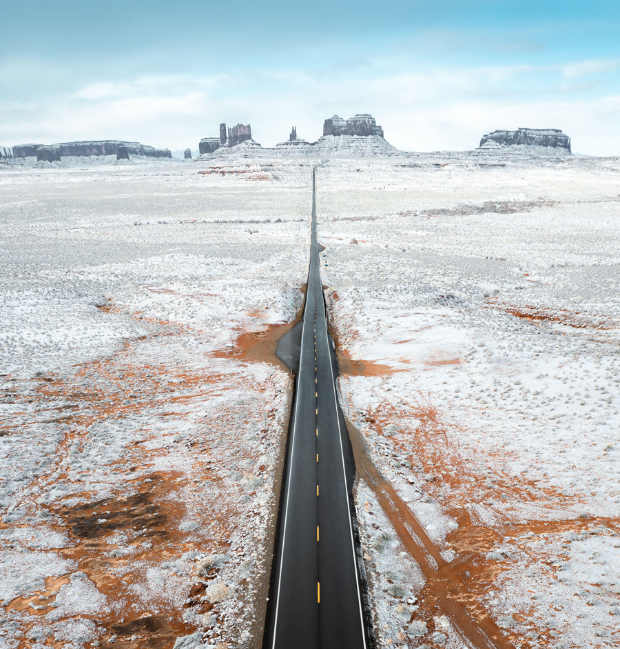 Aerial view of a stark black road cuts through the snow-dusted ochre earth towards the distant Monument Valley's sandstone buttes, Utah, United States