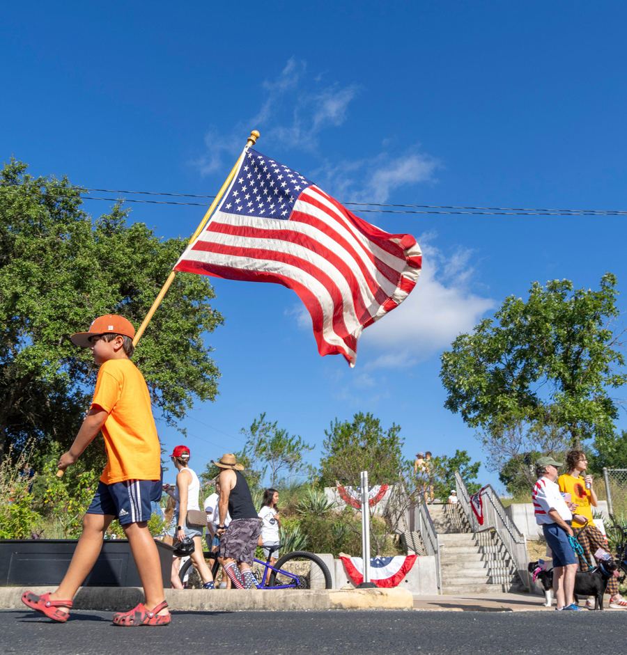 Ein Junge, der eine große amerikanische Flagge trägt, läuft am 4. Juli 2024 während der jährlichen Parade zum Unabhängigkeitstag im Stadtteil Barton Hills im Süden von Austin den Barton Hills Drive entlang.