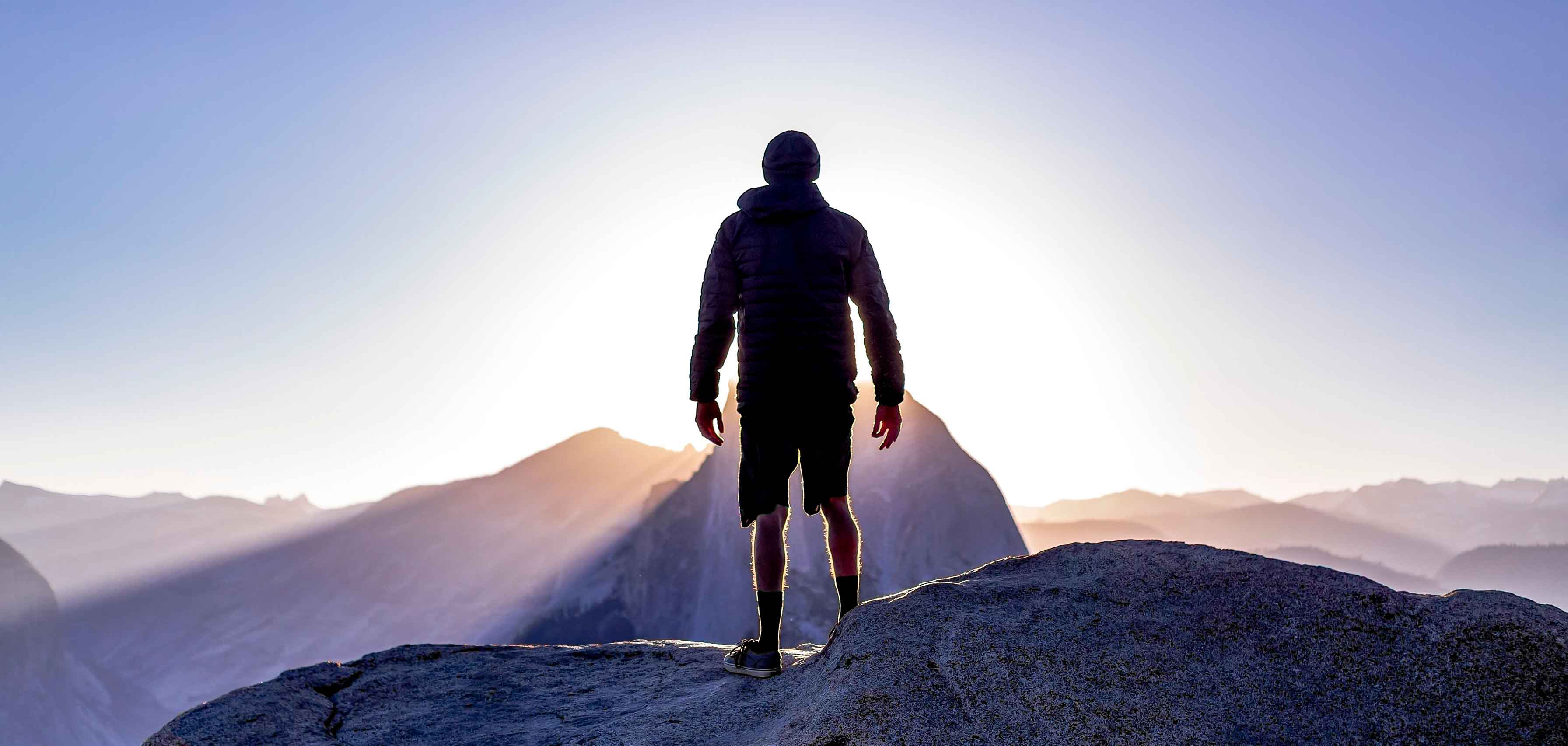 Mann auf einer Klippe mit Blick auf die Sonne und die Berge