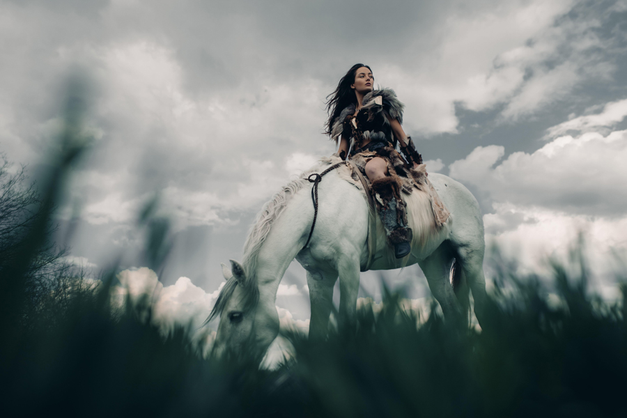 Woman rides on horseback in field in image of ancient warrior amazon on sky background.