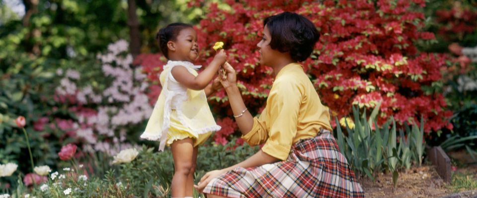 1980s SMILING AFRICAN AMERICAN WOMAN AND LITTLE GIRL OUTDOORS NEAR SPRING FLOWERS DAUGHTER GIVING YELLOW FLOWER TO MOTHER