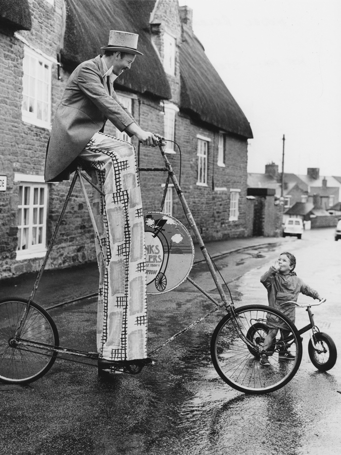 Man on stilts with a curious big bike looking down at a small boy, England, Great Britain