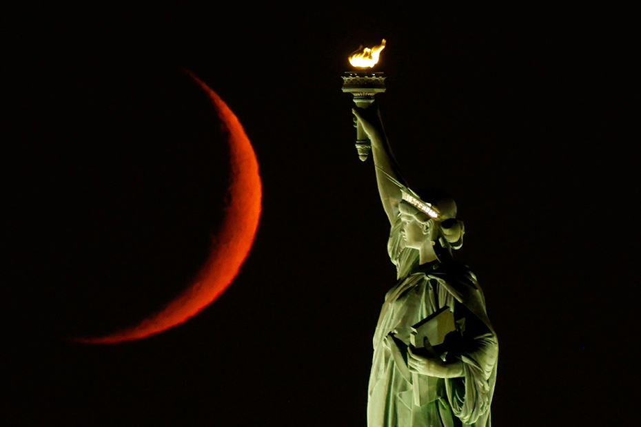 A waxing crescent moon sets behind the Statue Of Liberty in New York City