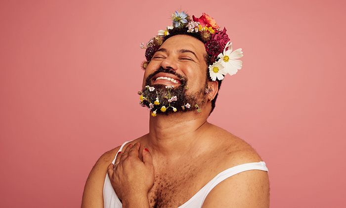 Gender queer smiling on pink background. Gay man wearing makeup and flowers on head and beard with copy space. 