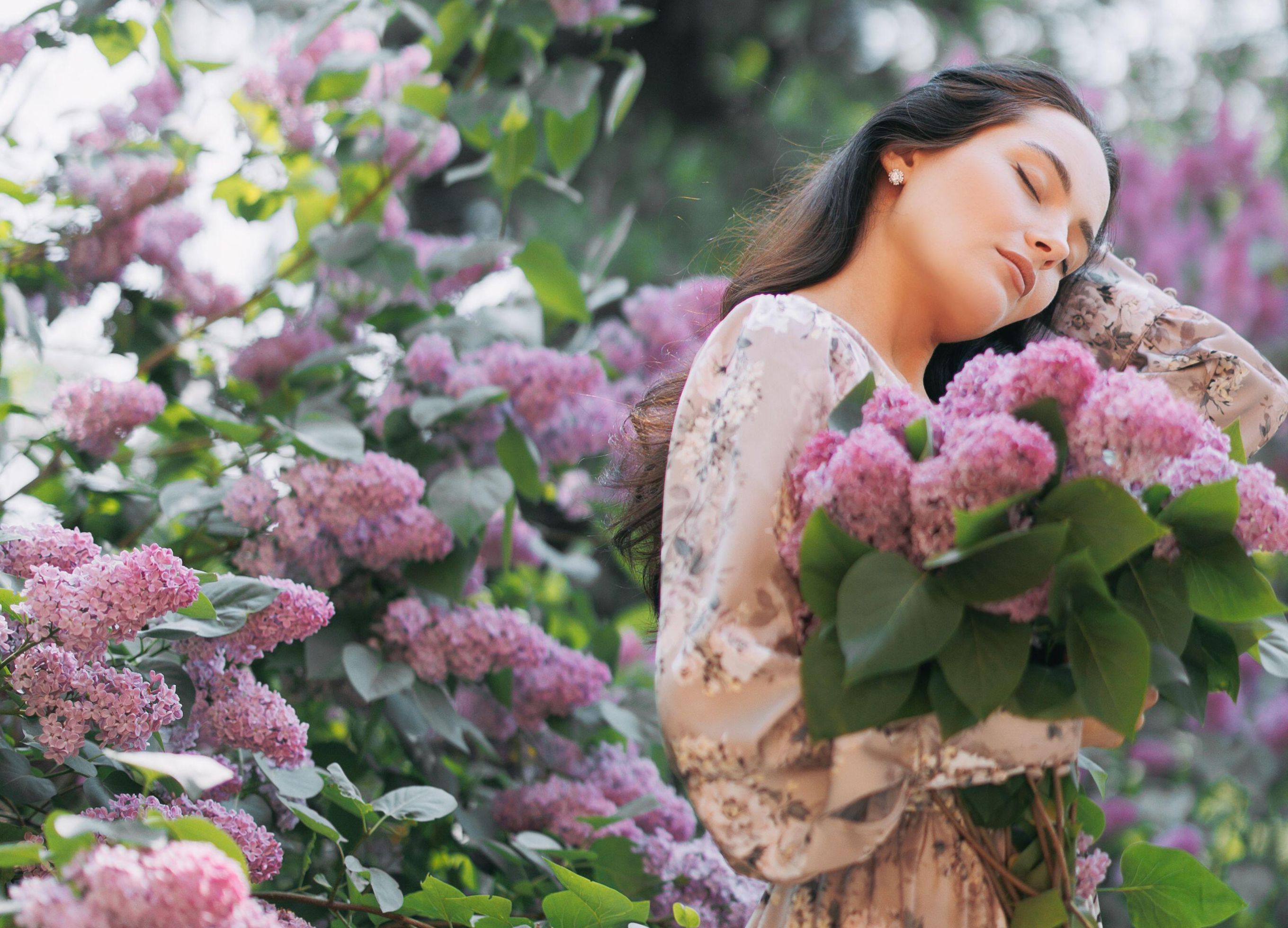 Una giovane e bella donna bruna cammina nel giardino con un mazzo di fiori in mano e si gode con gli occhi chiusi i cespugli di lillà in fiore. - ID dell'immagine: 2R1RCNP (RF)